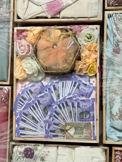 A creative way to present cash gifts as part of the wedding trousseau. The currency is artfully arranged in a pinewood tray, surrounded by other beautifully packed gifts and floral decorations.