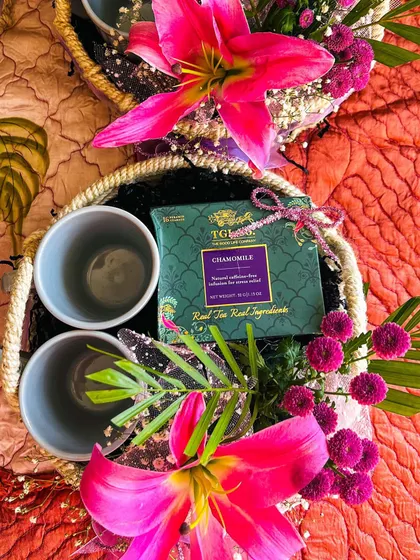 A vibrant floral hamper designed as a wedding or corporate favour. This basket features two ceramic mugs and a box of chamomile tea, surrounded by bright pink lilies and other colourful flowers, making it a refreshing and beautiful gift.
