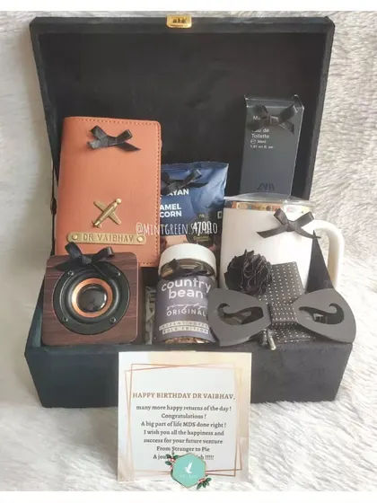 A birthday hamper for a doctor, personalized with his name in gold on a black suede box. Inside, a mix of useful items like a personalized passport cover, a Bluetooth speaker, and coffee.