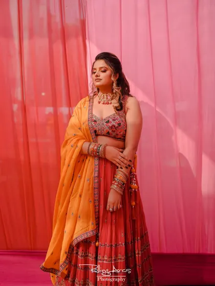 A full-length portrait of the bride at her Mehendi ceremony, showcasing her beautiful red and orange lehenga against a soft pink backdrop.