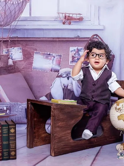 A little explorer ready for an adventure. This toddler, dressed in a vest and glasses, looks thoughtful as he sits at a desk with a globe and books in a vintage travel-themed shoot.