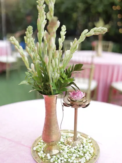 An old-world charm centerpiece with tuberoses in a brass vase and a lotus-shaped candle holder, set on a bed of jasmine flowers.