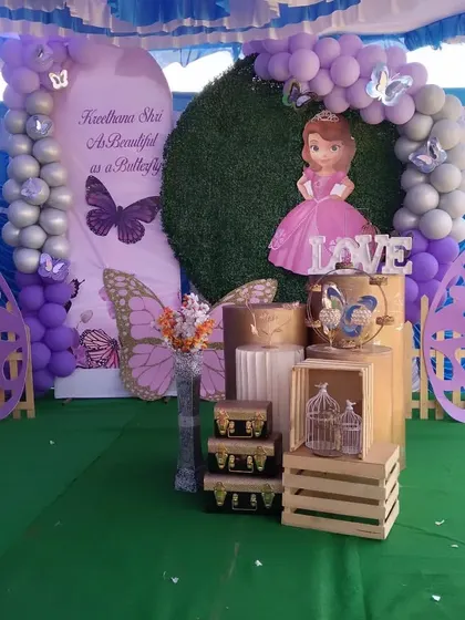 A beautiful butterfly and princess-themed setup for an outdoor party. This decor includes a grass wall, purple and silver balloon arch, and cutouts of butterflies and Princess Sofia.