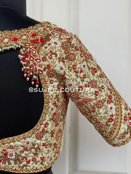 The back of the off-white wedding procession blouse. It features a deep, wide cutout with a pearl tassel detail at the top, framed by intricate floral and traditional motif embroidery.