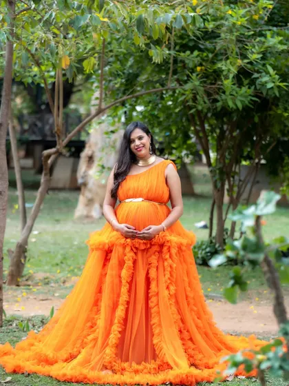 A full-length portrait of a mom-to-be in a vibrant orange gown, surrounded by the greenery of a park.