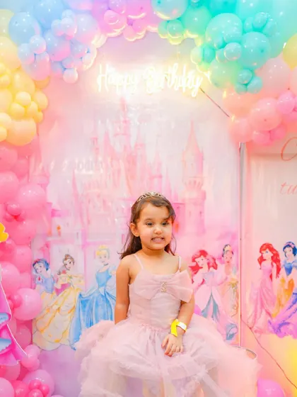 A beautiful princess-themed birthday party, with a backdrop of a pink castle and famous Disney princesses. The birthday girl is dressed perfectly for her royal celebration.