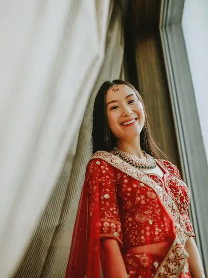 A happy, natural portrait of a guest or bridesmaid in a beautiful red lehenga, smiling by the window.