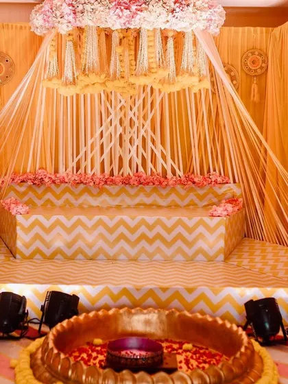 Here is the full view of the beautiful Haldi ceremony stage I designed. The setup features a canopy of hanging floral strings, a tiered seating area with a chevron pattern, and a large brass uruli in the front for the rituals.