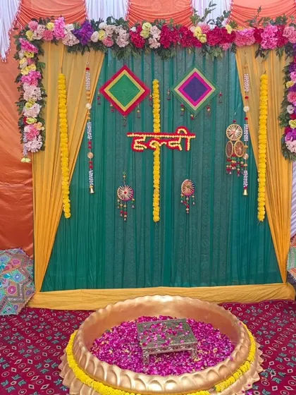 A traditional Haldi ceremony setup with a large brass urli filled with rose petals. The backdrop features green and yellow drapes, marigold garlands, and colorful hangings.
