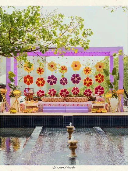 The Haldi stage set against a water feature, with a backdrop of giant floral lollipops and a canopy of lilac drapes, creating a stunning and unique ceremony space.