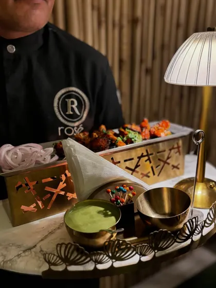 A server presents a tray of assorted appetizers, including chicken tikka with green chutney. The combination of marble, brass, and fresh ingredients creates a luxurious and appealing presentation.