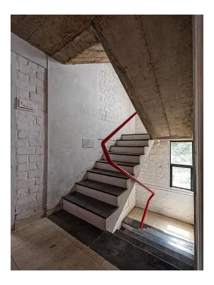 The common staircase in the 'Stacked Student Housing' building. The design uses simple, robust materials like concrete and stone, with a slender red handrail adding a line of color that guides movement through the building.