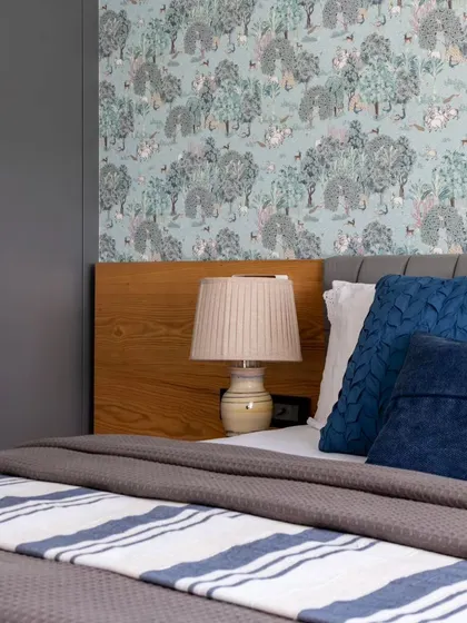 A close-up of the bedside in the Jaipur penthouse, showing the detailed wallpaper and the contrast between the grey upholstered headboard and the warm wooden side panel. A classic lamp adds a touch of timeless charm.