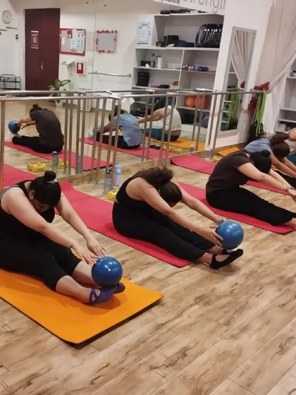A group of women in a mat class perform a forward fold stretch using Pilates balls to deepen the release in their hamstrings and lower back.