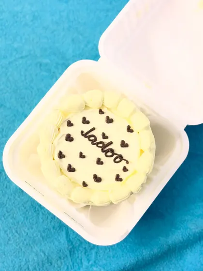 A simple and sweet bento cake with "Ladoo" written on it, surrounded by tiny chocolate hearts. The pale yellow frosting and piped border give it a classic, adorable look.