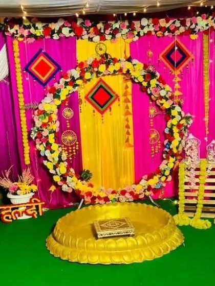 A vibrant Haldi ceremony setup with a circular floral arch against a backdrop of pink and yellow drapes. The decor includes a golden 'urli' and traditional hanging elements.