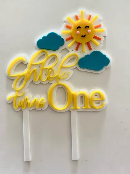 "Shlok turns One." This layered acrylic topper combines a cheerful yellow with a clean white font and a cute sun detail, making it a bright and happy addition to a first birthday cake.