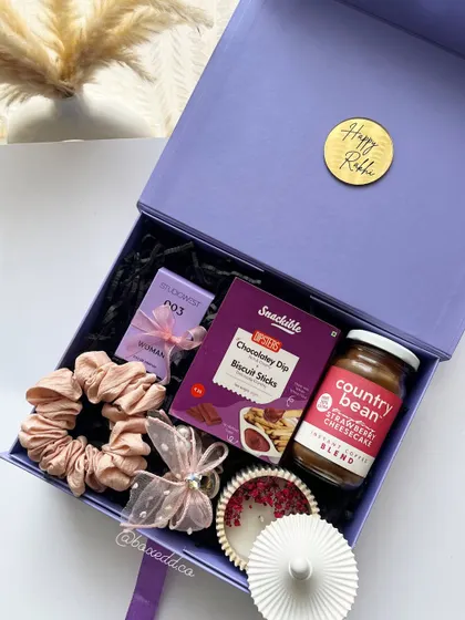 A lovely purple-themed Rakhi gift box for a sister, containing a scented candle, coffee, snacks, and scrunchies.