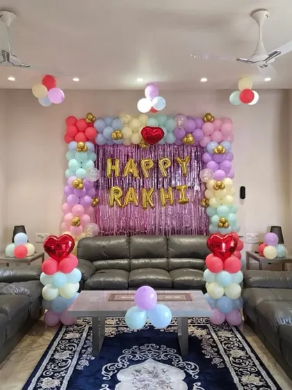 A festive decoration for a Rakhi celebration at home. The living room is decorated with a pastel-colored balloon frame and 'Happy Rakhi' foil letters.