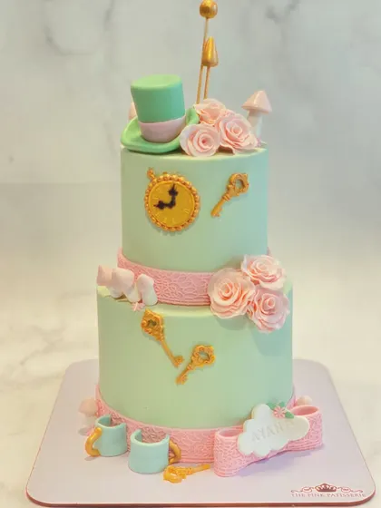 An "Alice in Wonderland" themed two-tiered cake. This whimsical creation includes iconic elements like the Mad Hatter's hat, a clock, keys, and teacups, all handcrafted from fondant.