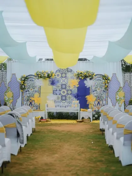 An outdoor Haldi setup with a Mediterranean vibe, featuring blue and yellow tile patterns, light fabric drapes, and guest seating arranged for the ceremony.