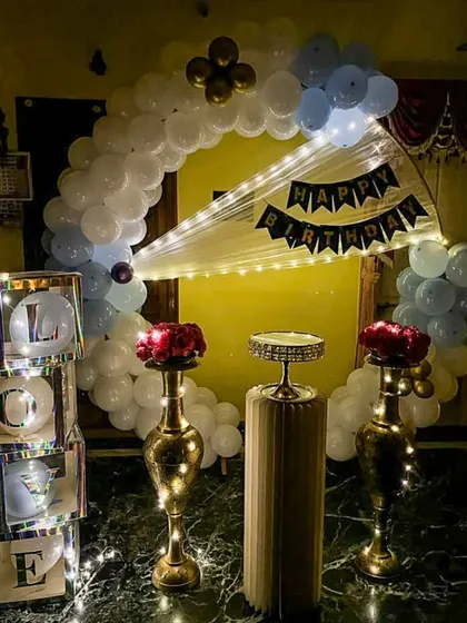 We create loving tributes for your special occasions. This home setup features a blue and white balloon arch, elegant brass vases with red roses, and illuminated "LOVE" display boxes for a heartfelt anniversary or birthday celebration.
