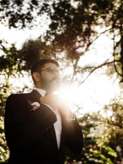 A groom adjusting his tuxedo, backlit by the sun through the trees. This shot has a classic, cinematic feel, capturing a moment of quiet preparation.