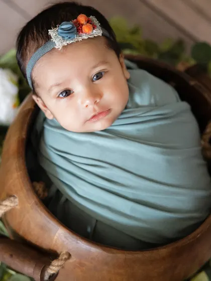 A close-up of a newborn's beautiful face, wrapped in a soft teal swaddle. The delicate headband adds a touch of detail to this simple and sweet portrait in a wooden bowl.
