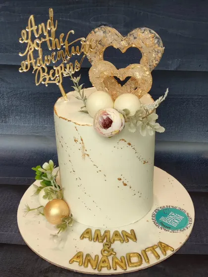 A simple and elegant cake for a couple's special day. The design features a clean white finish with gold leaf accents and a custom topper that reads "And so the Adventure Begins."