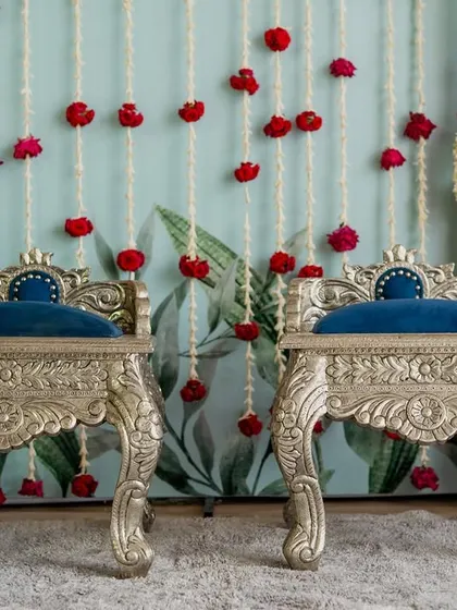 Ornate silver chairs with deep blue cushions, set for a traditional ceremony against a backdrop of hanging red and white flowers.