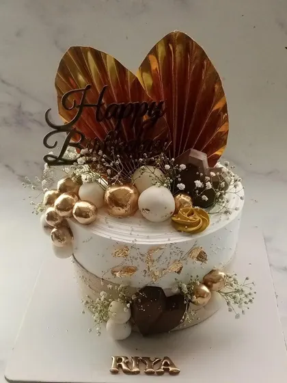 A stunning cake in white and gold, perfect for an elegant celebration. The design features gold-dusted palm leaves, chocolate spheres, and delicate baby's breath for a touch of luxury.