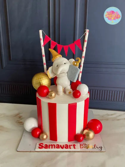 A charming circus cake with a hand-sculpted fondant elephant, festive red and white stripes, and playful gold and red spheres.