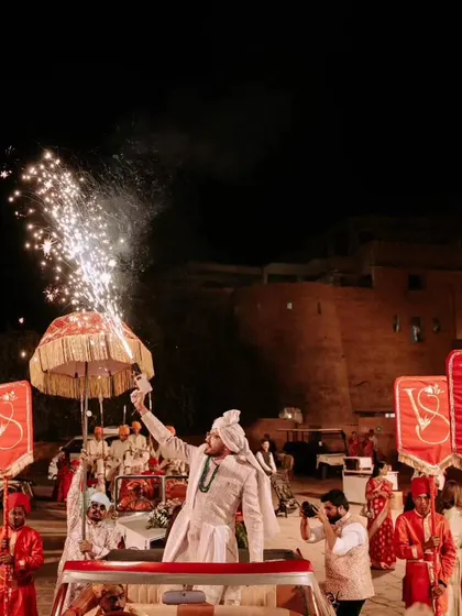 The grand Baraat entry at Suryagarh. The groom arrives in a vintage car, holding a sparkler, with monogrammed royal banners leading the procession.
