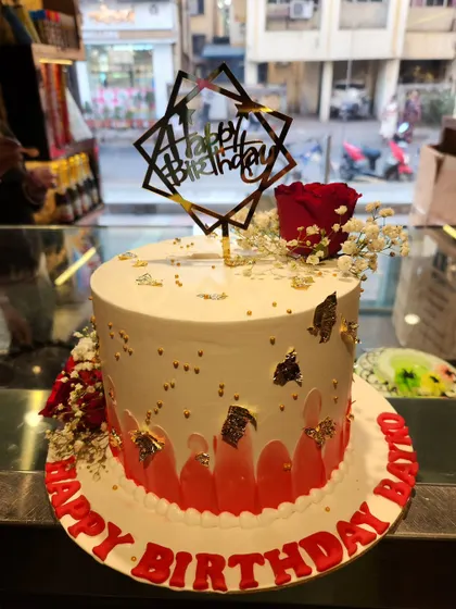 A chic and celebratory birthday cake. This design combines a red watercolor effect with gold leaf accents, fresh flowers, and a modern geometric 'Happy Birthday' topper.