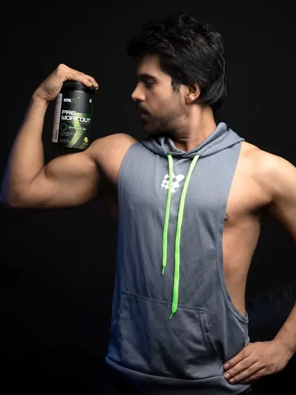 A male model in a sleeveless hoodie showing off his physique while holding a pre-workout supplement, conveying energy and readiness.