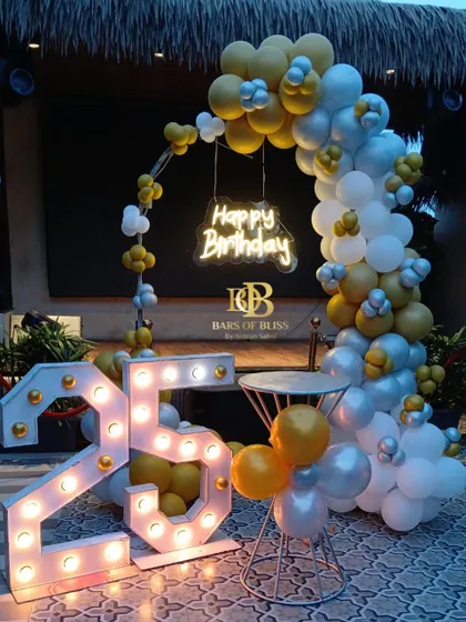 An outdoor 25th birthday celebration with a yellow, silver, and white color scheme. The decor features a balloon arch, a 'Happy Birthday' neon sign, and large marquee numbers that stand out beautifully against the evening sky.