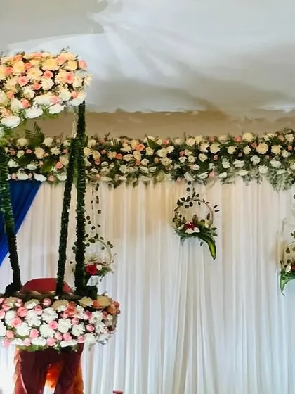 A grand naming ceremony setup with a hanging cradle decorated with pink and white flowers. The floral arch and blue and white curtains create a dreamy and celebratory atmosphere.