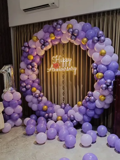 A beautiful circular balloon arch in shades of purple for a first anniversary. The setup is completed with a "Happy Anniversary" neon sign in the center.