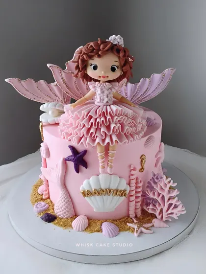 A beautiful mermaid-themed cake in shades of pink and lavender. The cake is adorned with a handcrafted mermaid figurine, seashells, and other underwater details.