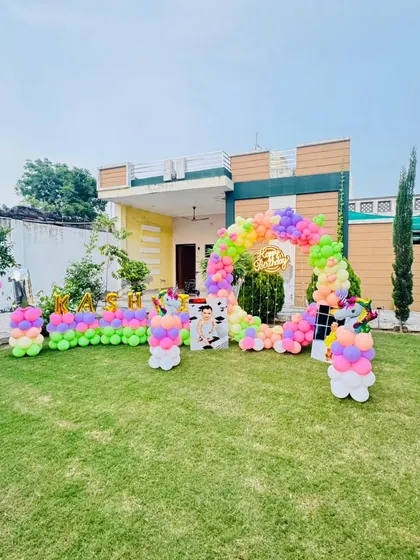 An outdoor unicorn-themed birthday party setup on a lush green lawn. The decor includes a pastel balloon wall with the name "KASHVI", a circular arch, and unicorn figures.