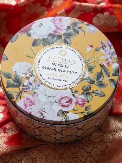 Our Marsala Geranium & Musk three-wick tin candle, resting on a rich, red silk fabric. The floral design and warm, musky scent make it a perfect gift to send to loved ones.