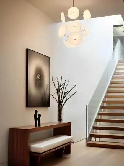 A bright and modern entryway next to a staircase. The space is furnished with a simple bench, a large piece of abstract art, and a contemporary chandelier.