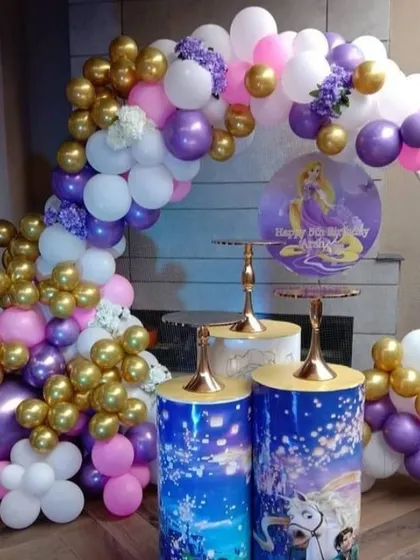 A Rapunzel-themed birthday party with character cutouts on themed plinths. The balloon arch uses purple, pink, and gold balloons to match the theme.