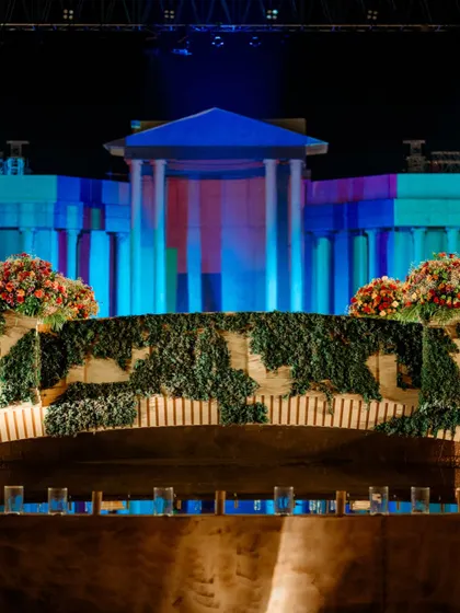 The Roman bridge, covered in ivy, with the grand stage illuminated by colorful lights in the background. This view highlights the different zones and architectural elements created for the event.