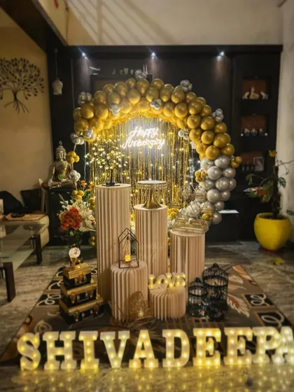 An elegant anniversary setup in a living room, featuring a gold and silver balloon arch, a 'Happy Anniversary' neon sign, and my signature prop display.