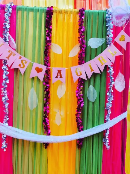 A close-up of a backdrop for a baby girl's welcome party. The 'It's A Girl' banner is hung against a multi-colored drape, showing a simple yet sweet way to announce your joy.