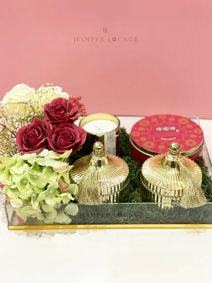 This acrylic tray hamper is filled with festive red and gold items for Eid. It includes decorative jars with tassels, a scented candle, and a tin of sweets, all nestled on a bed of green moss for a vibrant look.