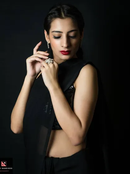 An elegant low-key portrait of a woman in a black saree. The lighting gently illuminates her face and hands, highlighting her red lipstick and diamond jewelry against the dark background.