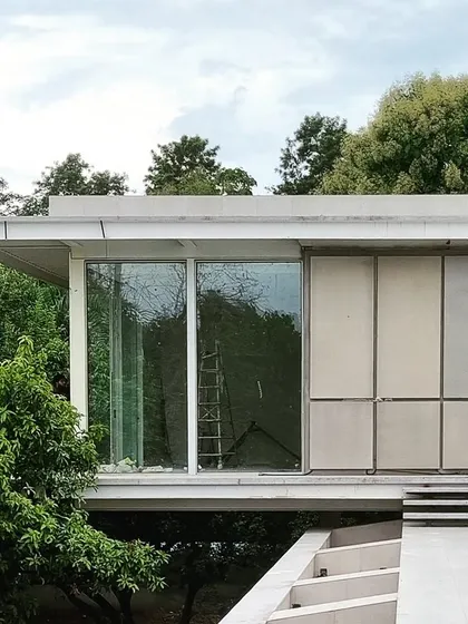 A house designed as a pavilion floating above a garden. The structure features large glass panels and a handcrafted metal skin with a soft patina, designed to blur the boundaries between inside and out and respond to the changing light.