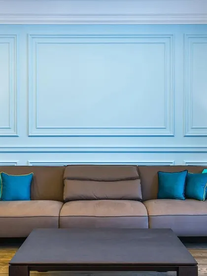 A close-up of the living area, showing a comfortable brown sofa accented with bright blue cushions, set against a powder-blue wall with classic decorative molding.